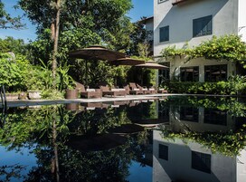 Uma área de piscina tranquila com espreguiçadeiras e guarda-sóis, rodeada por vegetação exuberante e um edifício branco com telhado em azulejos.