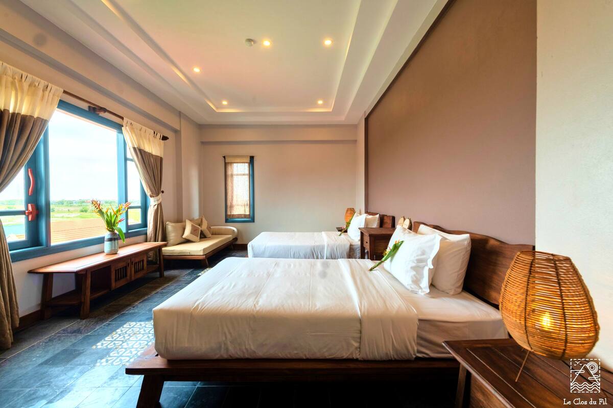 A hotel room with two beds, a bench, a window with curtains, a bedside table with a lamp, and a view of the outdoors.