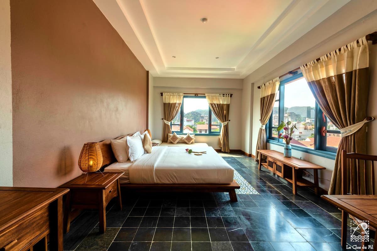 A hotel room with a large bed, a wooden bench, a small table, and a view of the city through the windows.