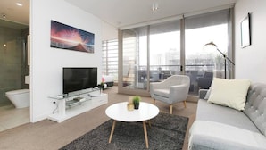 A modern living room with a flat-screen TV, a round coffee table, a grey sofa, and a large window offering a city view. - HUGO - Live the good life *Wifi GYM Parking Views (Melbourne)