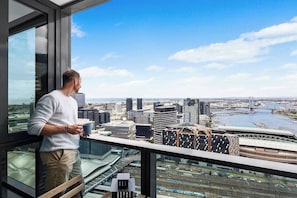 A man standing by a window overlooking a cityscape with modern buildings and a stadium.