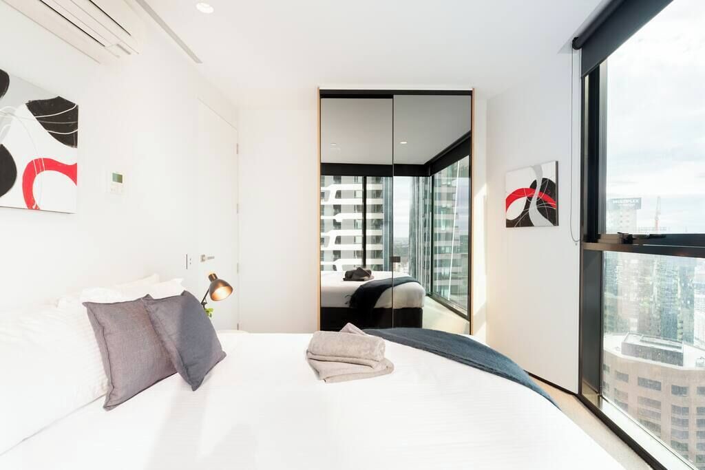 Ein modernes Hotelzimmer mit einem großen Bett, einem Nachttisch mit Lampe, Blick auf die Stadt durch große Fenster und einer Schiebetür zu einem Balkon.