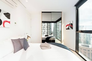 Een moderne hotelkamer met een groot bed, een nachtkastje met lamp, uitzicht op de stad door grote ramen en een schuifdeur naar een balkon.