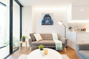 Een moderne woonkamer met een grijze bank, een ronde salontafel met een plant, een schilderij aan de muur, en een keuken met een spoelbak en kastjes.