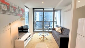 A modern living room with a flat-screen TV, a glass coffee table, a sofa, and a large window with a view of buildings. - Phoenix - Amazing Location with Gym Sauna Pool Internet (Melbourne)