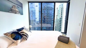 A hotel room with a large bed, a pillow, a bedside lamp, a small rug, and a view of tall buildings through a large window. - Phoenix - Amazing Location with Gym Sauna Pool Internet (Melbourne)