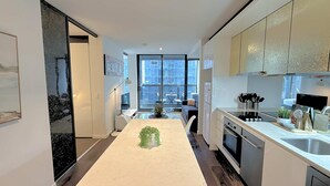 A modern kitchen with a central island, a dining area with chairs, and a large window offering a city view. - Phoenix - Amazing Location with Gym Sauna Pool Internet (Melbourne)