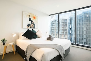 Une chambre moderne avec un grand lit, une table de chevet, une lampe, un tableau et une vue sur les immeubles à travers de grandes fenêtres.