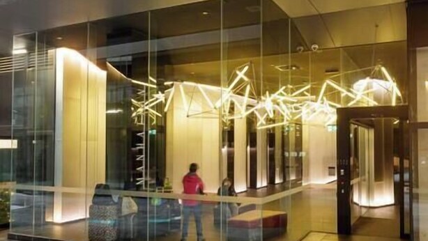 A modern building entrance with glass walls, a geometric light fixture, and a person standing in the lobby.