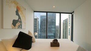 A hotel room with a large bed, a window with a city view, a painting on the wall, and a sign indicating the hotel name.