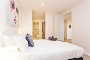 A bedroom with a bed, pillows, a bedside lamp, a door leading to a bathroom, and a hallway.