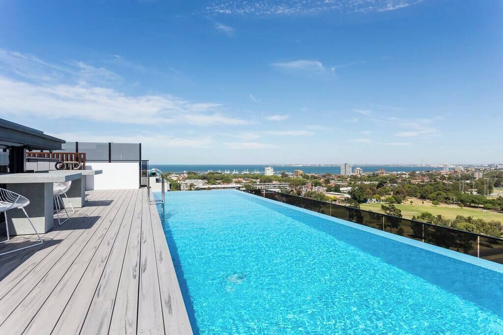 Ein Infinity-Pool auf dem Dach mit Holzterrasse, Sitzgelegenheiten im Freien und Blick auf die Stadt.