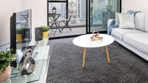A modern living room with a glass coffee table, a white sofa, and a large window with a city view. - Orianna - Stylish Designer Pad *WiFi Park Gym Pool (South Yarra)