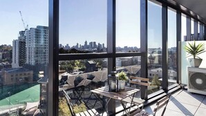 A high-rise room with large windows, a view of the city skyline, a small table with a plant, and a potted plant on a stand. - Orianna - Stylish Designer Pad *WiFi Park Gym Pool (South Yarra)