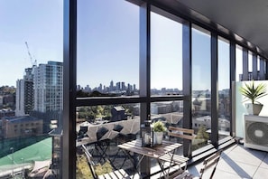 A high-rise room with large windows, a view of the city skyline, a small table with a plant, and a potted plant on a stand.