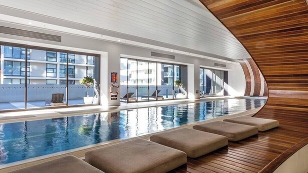 An indoor swimming pool with a wooden deck, large windows, and lounge chairs. - Orianna - Stylish Designer Pad *WiFi Park Gym Pool (South Yarra)