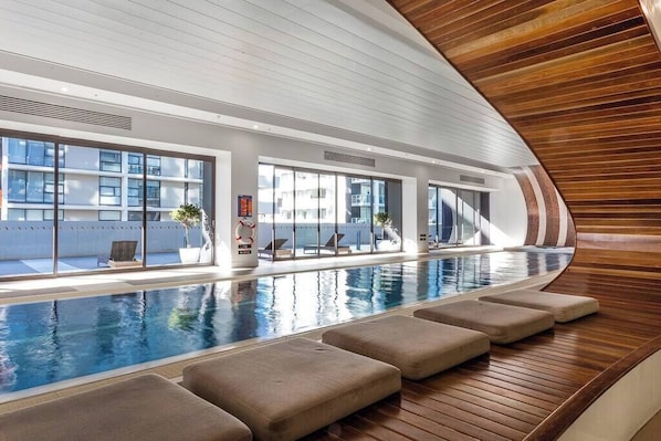 An indoor swimming pool with a wooden deck, large windows, and lounge chairs.