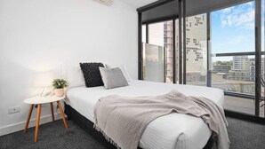 A modern bedroom with a large bed, a small bedside table with a lamp, a potted plant, and a view of buildings through floor-to-ceiling windows. - Orianna - Stylish Designer Pad *WiFi Park Gym Pool (South Yarra)