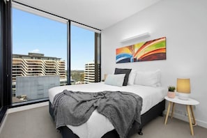 A modern bedroom with a large bed, a view of buildings, and a colorful abstract painting on the wall.