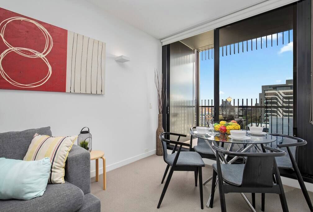 A modern living room with a grey sofa, a glass dining table, and a large window with a city view.