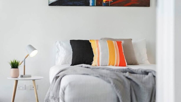 Une chambre bien rangée, avec un lit, une lampe de chevet et une petite table sur laquelle est posée une plante.