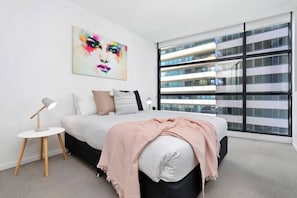 A modern bedroom with a large bed, a bedside table with a lamp, a colorful abstract painting on the wall, and a large window with a view of a building.