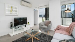 Een moderne woonkamer met een flatscreen televisie, een glazen salontafel, een grijze bank en een groot raam met uitzicht op de stad.