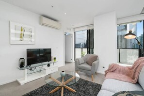 Un soggiorno moderno con televisore a schermo piatto, tavolino in vetro, divano grigio e un'ampia finestra con vista sulla città.