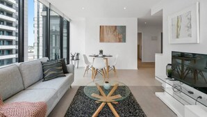 Een moderne woonkamer met een bank, een glazen salontafel, een eethoek met stoelen, een televisie en kunstwerken aan de muur.