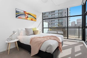 A modern bedroom with a large window, a bed with multiple pillows, a bedside table with a lamp, and a painting on the wall.