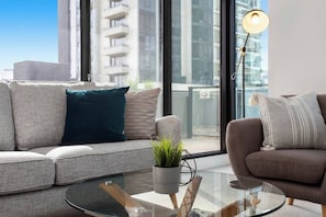A modern living room with a grey sofa, a round glass coffee table, and a view of a cityscape through large windows.