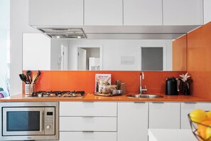 A modern kitchen with white cabinets, an orange backsplash, a stainless steel stove, a microwave, and a kettle.