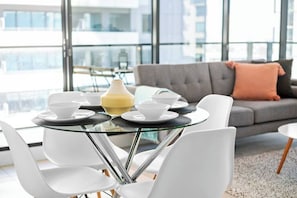 A modern dining area with a glass table, white chairs, and a grey sofa with a peach pillow.