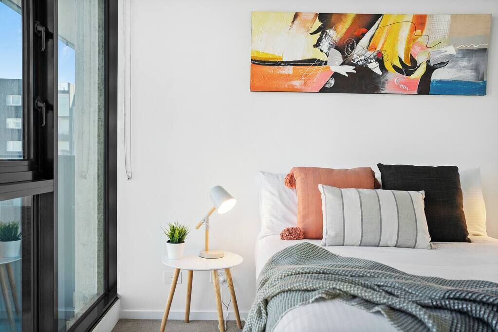 A bedroom with a bed, pillows, a bedside table with a lamp, a small plant, and a painting on the wall.