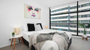 Ein modernes Schlafzimmer mit einem großen Bett, einem Fenster mit Stadtblick, einem kleinen Nachttisch und einem Wandbild.
