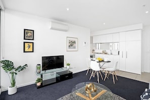 A modern living room with a flat-screen TV, a glass coffee table, and a dining area with a white table and chairs.