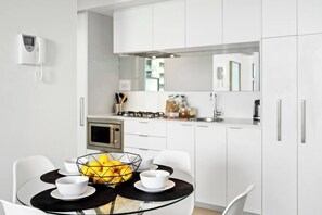 Una cocina moderna con armarios blancos, una mesa de vidrio para comer con cuatro cuencos blancos y un frutero.
