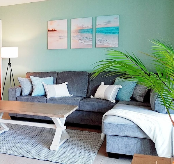 A living room with a grey sofa, a coffee table, and a large green plant. There are three framed pictures on the wall.