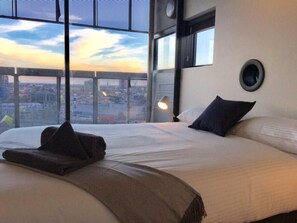 Habitación de hotel con una cama grande, una mesita de noche, una lámpara y vistas a un paisaje urbano a través de amplios ventanales.