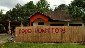 Una valla de bambú con la inscripción "DODO DORMITOR", un edificio de techo rojo y una persona parada al frente.