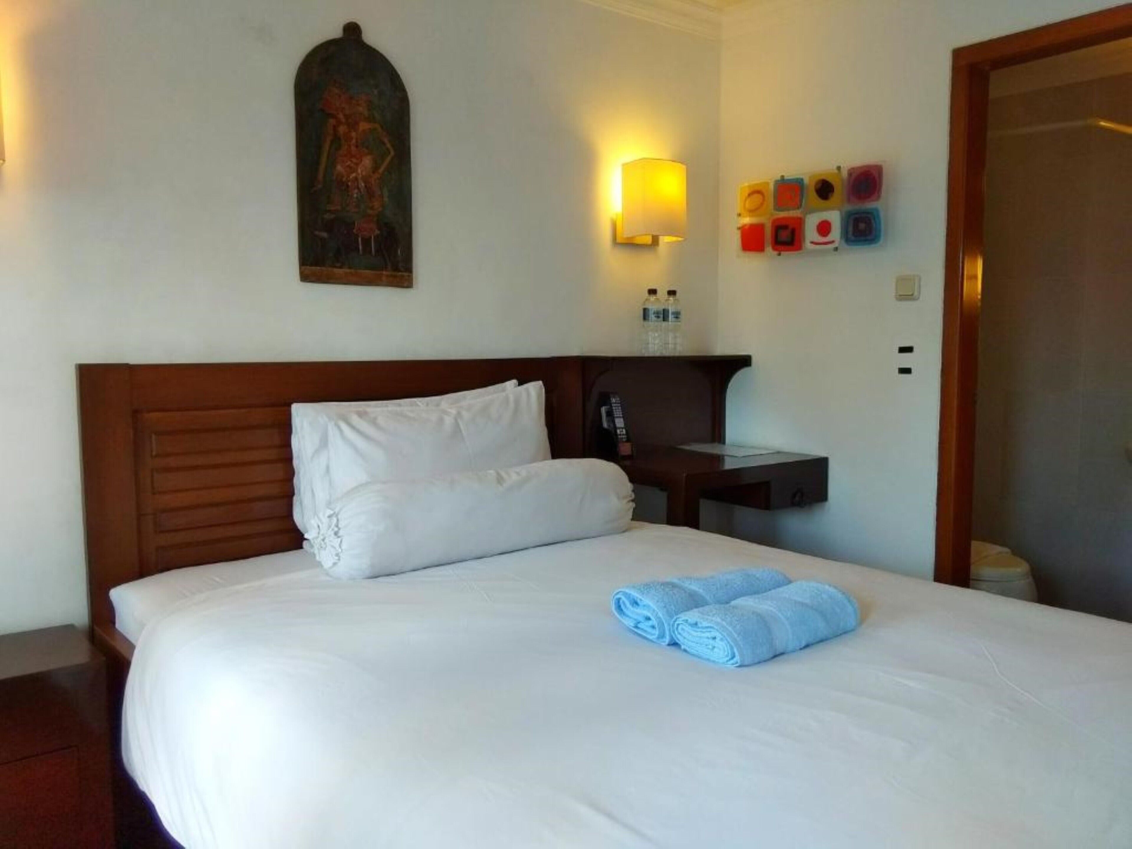 Habitación de hotel con cabecera de madera, ropa de cama blanca, un cuadro en la pared, una mesita de noche con lámpara y un teléfono en la pared.