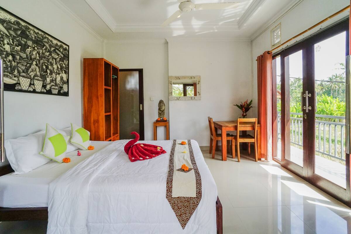 A well-lit room with a bed, a wooden cabinet, a small table, and a balcony with a view of greenery.