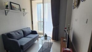 Ein modernes Wohnzimmer mit einem grauen Sofa, einem kleinen runden Tisch und einem Couchtisch. Es gibt ein Fenster mit Vorhängen, eine Pflanze an der Wand und eine kleine Ablage mit Dekorationen.