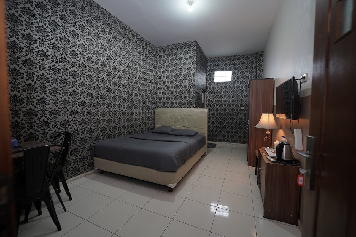 A bedroom with a bed, a desk, a chair, a TV, and a small window.