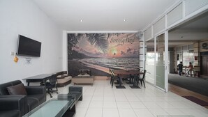 Un moderno vestíbulo de hotel con un gran mural de un atardecer playero, un televisor de pantalla plana y una zona de estar con mesas y sillas.