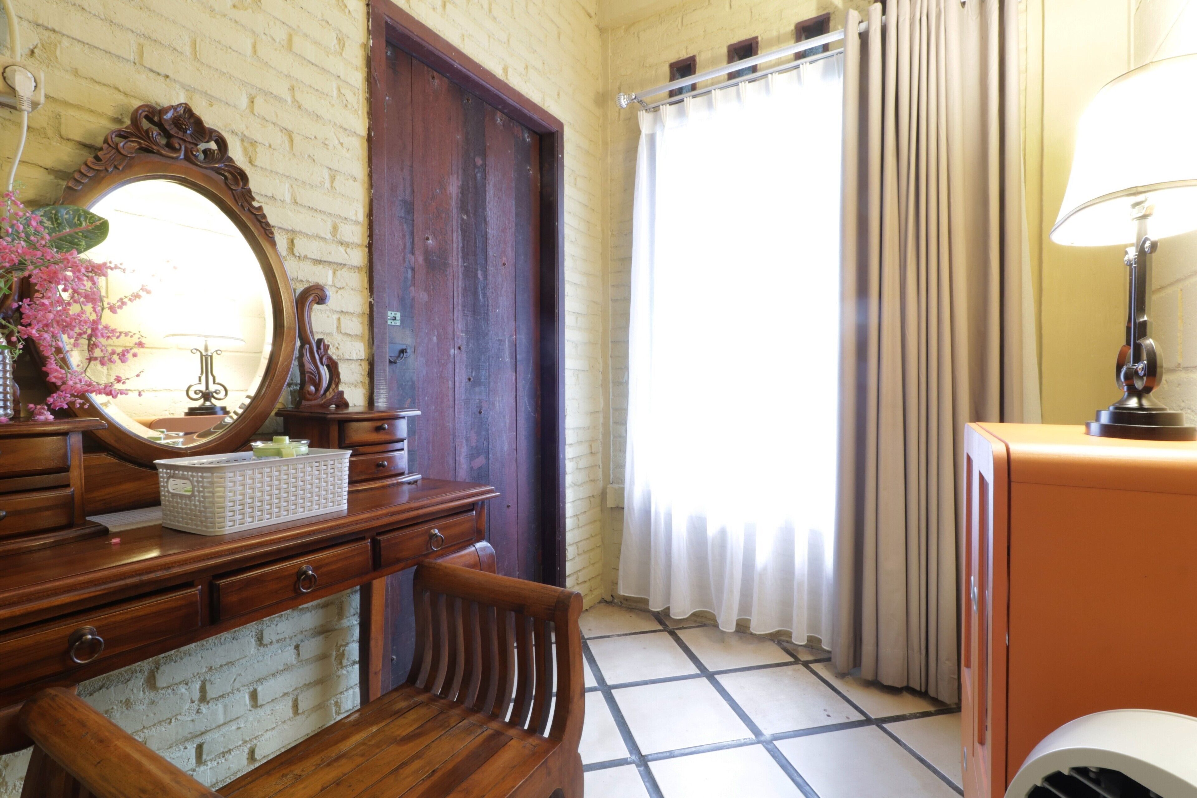 Um quarto com cômoda de madeira, espelho, cadeira, janela com cortinas e abajur.
