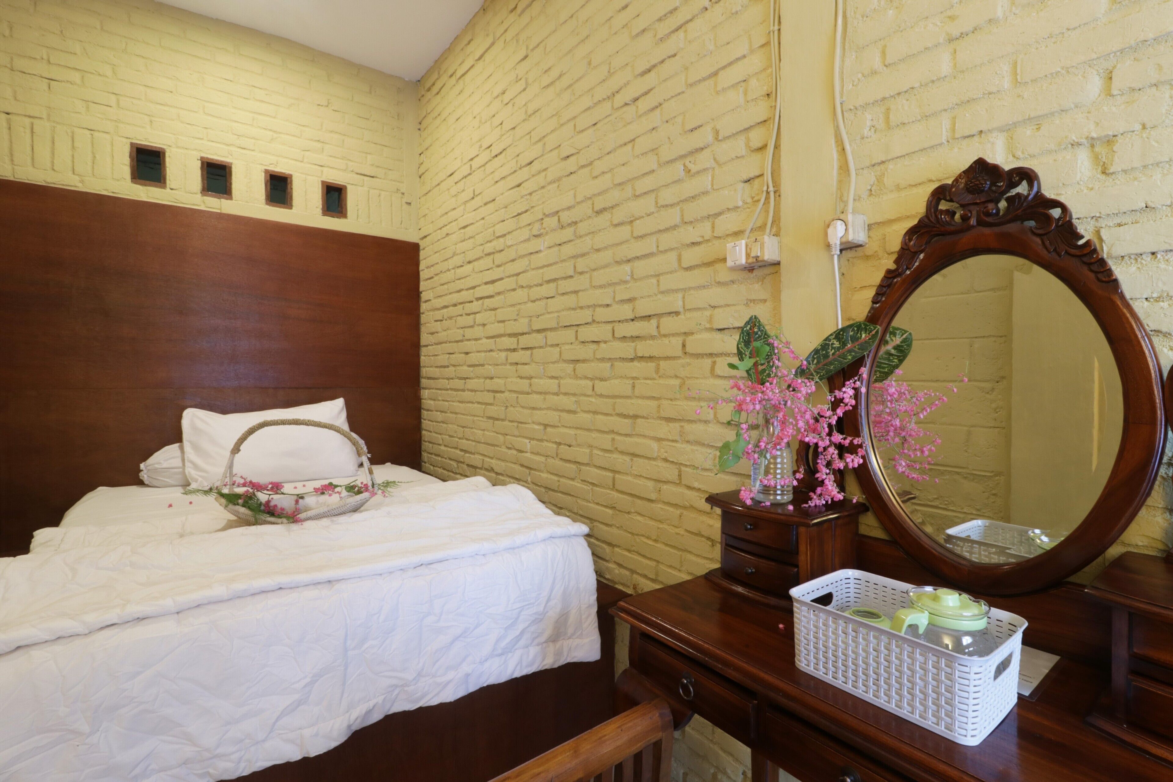 A hotel room with a brick wall, a wooden headboard, a bed with white bedding, a bedside table with a mirror, and a dresser.