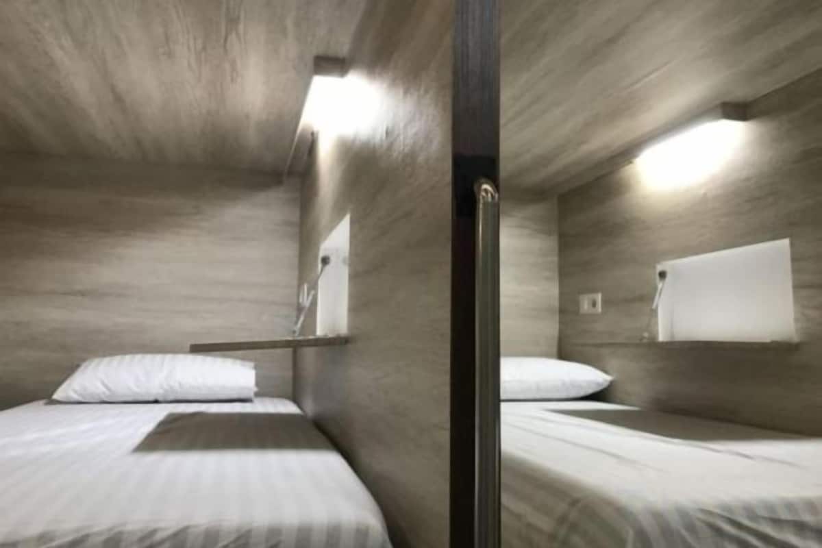 A compact room with two beds, a wooden wall, a small window, and a wall-mounted light fixture.