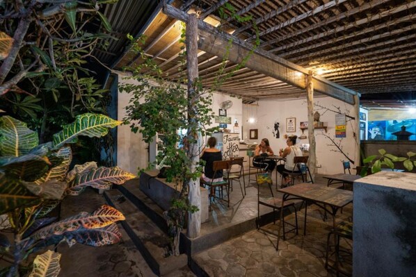 Un café en plein air, avec des tables et des chaises, entouré d’une végétation luxuriante et surmonté d’un plafond en bois rustique.