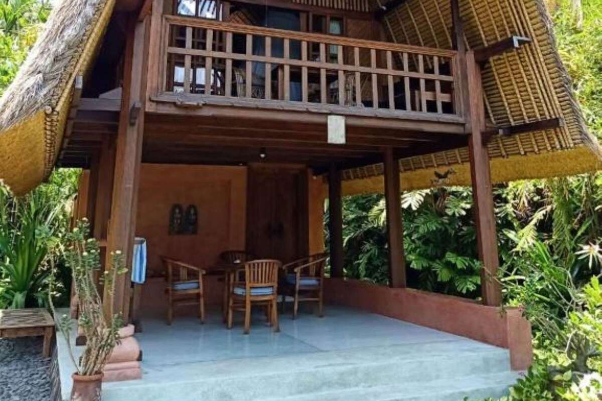 A two-story thatched-roof house with a balcony, surrounded by tropical vegetation.